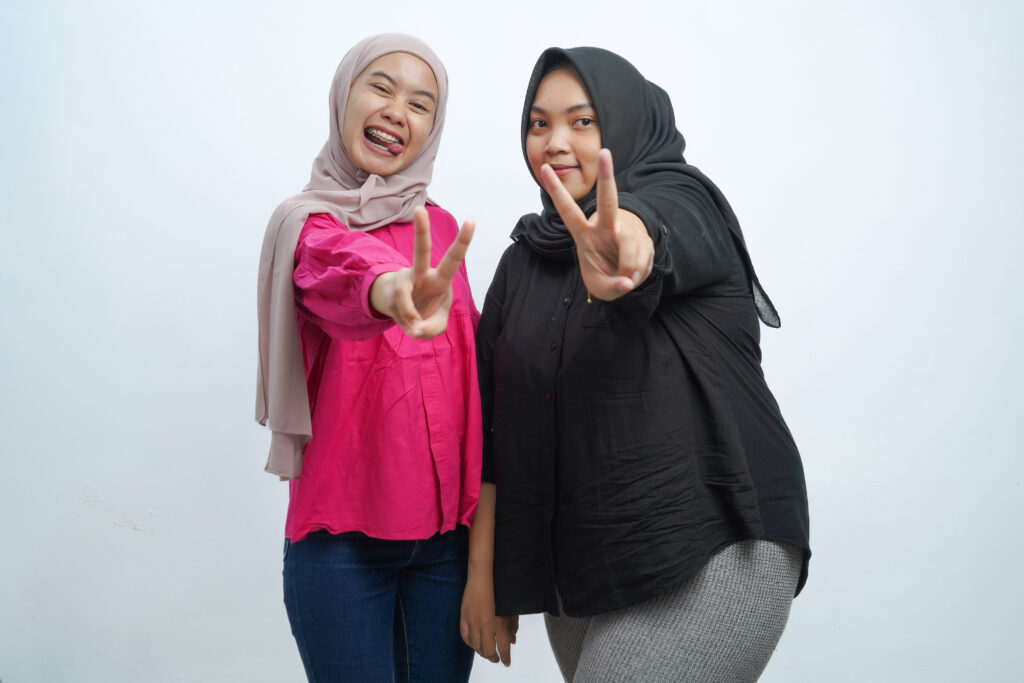 Two joyful Asian Indonesian Muslim women in hijabs making peace signs and smiling brightly while posing in a studio setting.